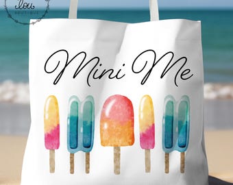 May include: White tote bag with the words "Mini Me" in black script. Below the text are five colorful watercolor popsicle illustrations. The bag has white straps and a logo in the upper left corner.