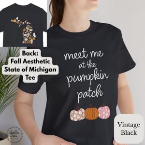 May include: Black t-shirt with the text "meet me at the pumpkin patch" in white script. Below the text are three pumpkins in different colours and patterns. The back of the shirt features a fall-themed Michigan state design.