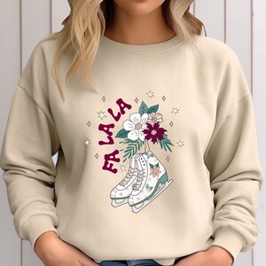 May include: A beige sweatshirt with a Christmas-themed graphic. The graphic features a pair of white ice skates with floral accents, surrounded by stars and the text "Fa La La" in burgundy.