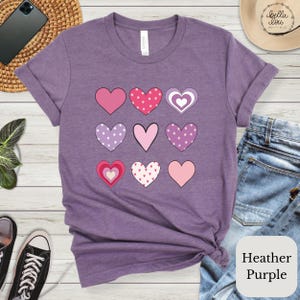 May include: A heather purple t-shirt with a design of nine pink, purple, and white hearts with different patterns. The shirt is laid out on a wooden surface with a pair of jeans and a small brown hat.