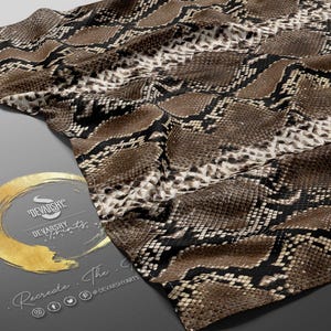 May include: A fabric with a snakeskin pattern in shades of brown, black, and cream. The material is draped, showing the texture and design. The image also includes a logo with gold accents.