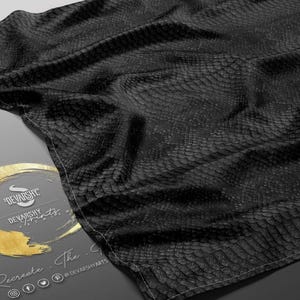 May include: A black fabric with a snakeskin pattern is draped over a surface. The fabric has a textured appearance and is folded. A gold and grey logo with the text "DEVANSHY PRINTS" is visible. The logo also includes social media icons.