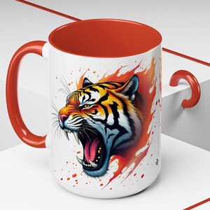 May include: A white ceramic mug with a red handle and rim. The mug features a colorful illustration of a tiger's head with orange, yellow, and black stripes. The tiger is roaring with its mouth open and teeth showing. The background is white with orange and yellow splatters of paint.