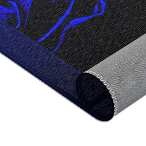 May include: A rectangular area rug featuring a black background with a vibrant blue abstract pattern. The rug is partially rolled, revealing a light gray textured underside and a dark blue bound edge that frames the top surface of the floor covering.