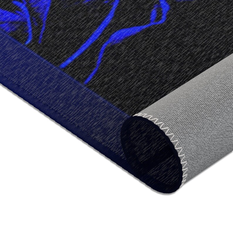 May include: A rectangular area rug featuring a black background with a vibrant blue abstract pattern. The rug is partially rolled, revealing a light gray textured underside and a dark blue bound edge that frames the top surface of the floor covering.
