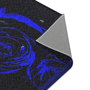 May include: A rectangular area rug featuring a dark charcoal gray background with a vibrant blue rose pattern. The corner of the rug is folded over to reveal a light gray underside with white stitched edges and a dark blue border trim.