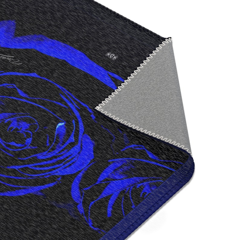 May include: A rectangular area rug featuring a dark charcoal gray background with a vibrant blue rose pattern. The corner of the rug is folded over to reveal a light gray underside with white stitched edges and a dark blue border trim.