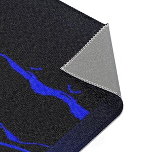 May include: A rectangular area rug featuring a dark charcoal gray textured surface with a vibrant blue abstract pattern. One corner of the rug is folded over to reveal a light gray underside with white stitched edges along the border.
