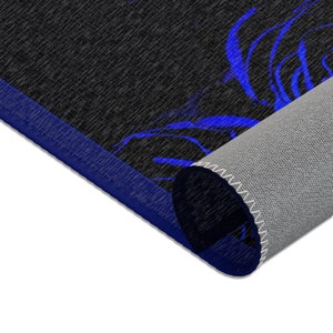 May include: A rectangular area rug featuring a black textured surface with a vibrant blue floral pattern. The rug has a solid blue border and is partially rolled, revealing a light gray, woven backing material with white stitched edges.