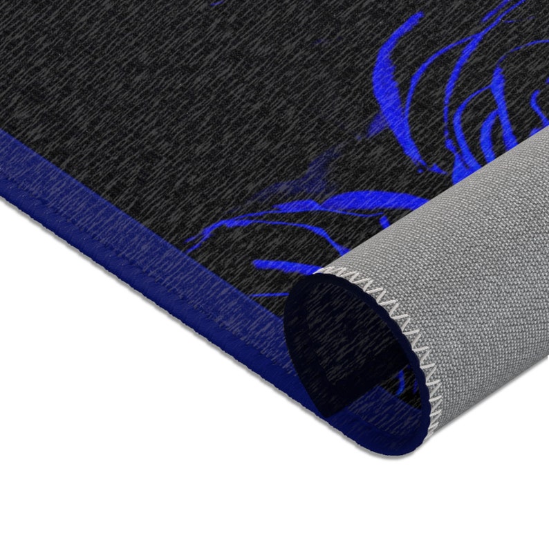 May include: A rectangular area rug featuring a black textured surface with a vibrant blue floral pattern. The rug has a solid blue border and is partially rolled, revealing a light gray, woven backing material with white stitched edges.