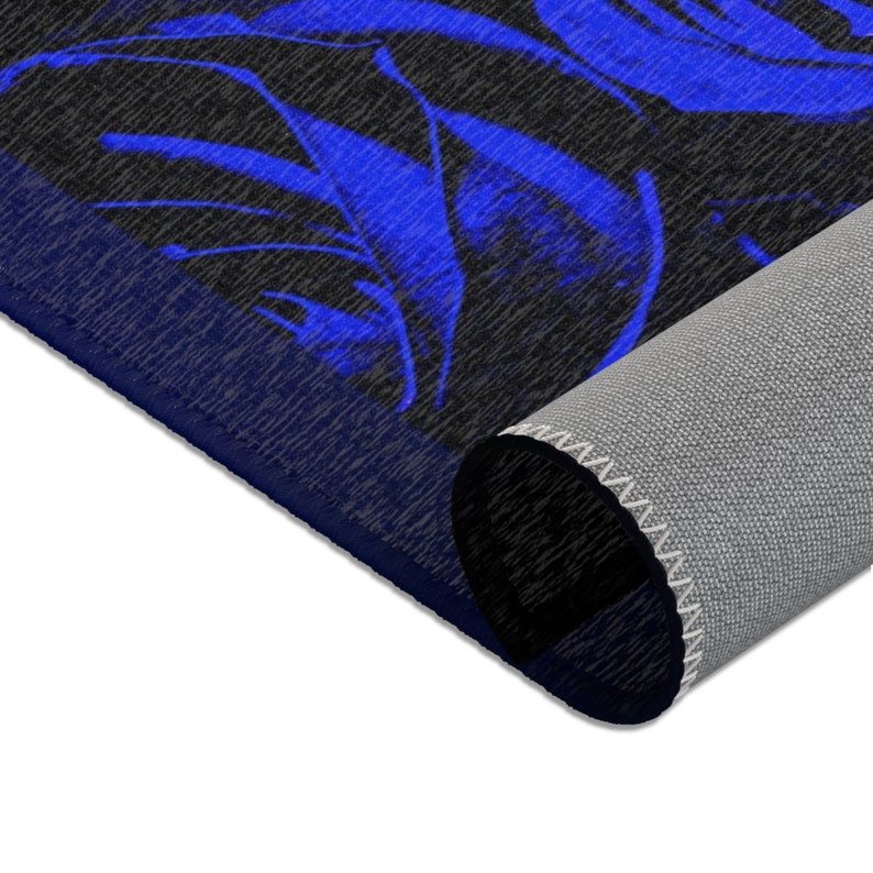 May include: A rectangular area rug featuring a bold, vibrant blue floral pattern set against a dark charcoal gray background. The rug is partially rolled, revealing a light gray, textured underside with a stitched edge along the border.