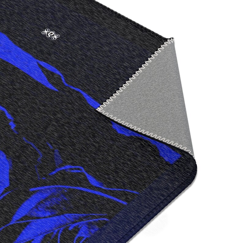 May include: A rectangular area rug featuring a dark charcoal gray heathered texture with bold, abstract royal blue brushstroke patterns. The corner is folded over to reveal a light gray underside with white serged edge stitching and a small logo.