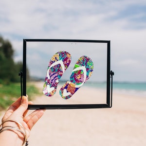 May include: A framed image of a pair of colorful flip-flops with a white background. The flip-flops are in a black frame and the image is being held in front of a beach scene.