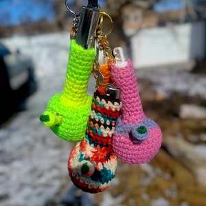 Op de afbeelding: Vier gebreide sleutelhangers in de vorm van een klein flesje. De sleutelhangers zijn in felle kleuren: groen, roze, rood en blauw. De sleutelhangers zijn bevestigd aan een zilveren sleutelhanger.