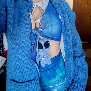 Könnte beinhalten: Ein blauer Strickcardigan mit Reißverschluss und einer großen Tasche auf der Vorderseite. Der Cardigan wird über einem blau-weiß gemusterten Oberteil und einem blau-weiß gemusterten Rock getragen. Ein kleines blaues Alien-Plüschtier hängt an einer Kettenkette.