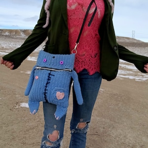Op de afbeelding: Een blauwe gehaakte monster-vormige schoudertas met een rits, knopen en een roze hartpatch. De tas wordt gedragen door iemand die een roze kanten top, een groene blazer en een blauwe jeans draagt.