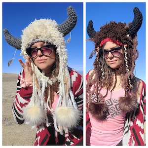 May include: Two women wearing crocheted buffalo hats with long, colorful yarn strands. The hats are white and brown with faux fur accents. The women are wearing colorful sweaters and tank tops.