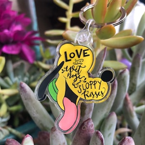 May include: A yellow and green cartoon dog keyring with a pink tongue sticking out. The keyring has the text "Love is a wet nose & sloppy kisses" on it.