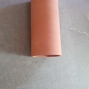 May include: A terracotta-colored cylindrical clay pipe, lying on a gray concrete surface.