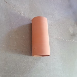 May include: A terracotta-colored cylindrical pipe, likely for drainage or ventilation, is shown against a gray concrete background.