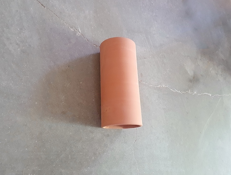 May include: A terracotta-colored cylindrical pipe, likely for drainage or ventilation, is shown against a gray concrete background.
