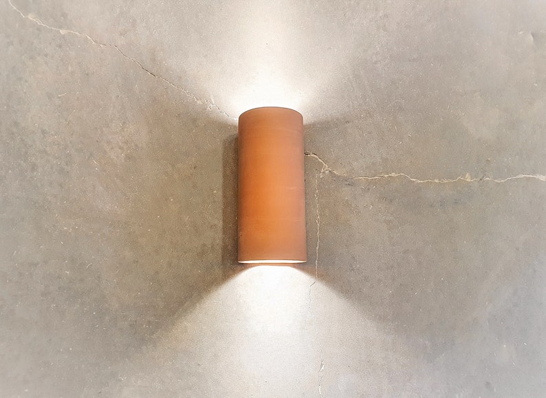 May include: A cylindrical terracotta wall sconce with a white light shining down on a concrete wall.
