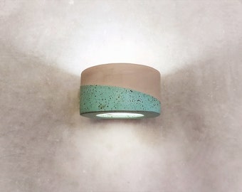 Puede incluir: Una lámpara de pared moderna hecha de hormigón con un acabado moteado verde claro y blanco. La lámpara tiene una forma circular y un agujero en el centro para una bombilla.