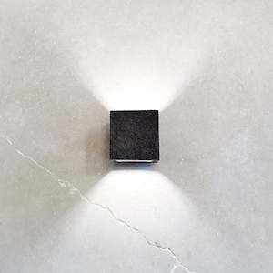 May include: A black square-shaped outdoor light fixture mounted on a gray wall. The light is on and casting a bright white light.
