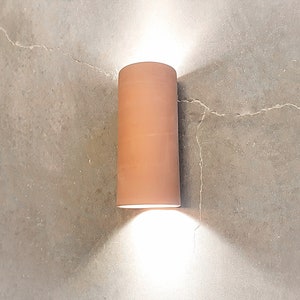 May include: A terracotta cylindrical wall sconce with a white light shining down on a concrete wall.