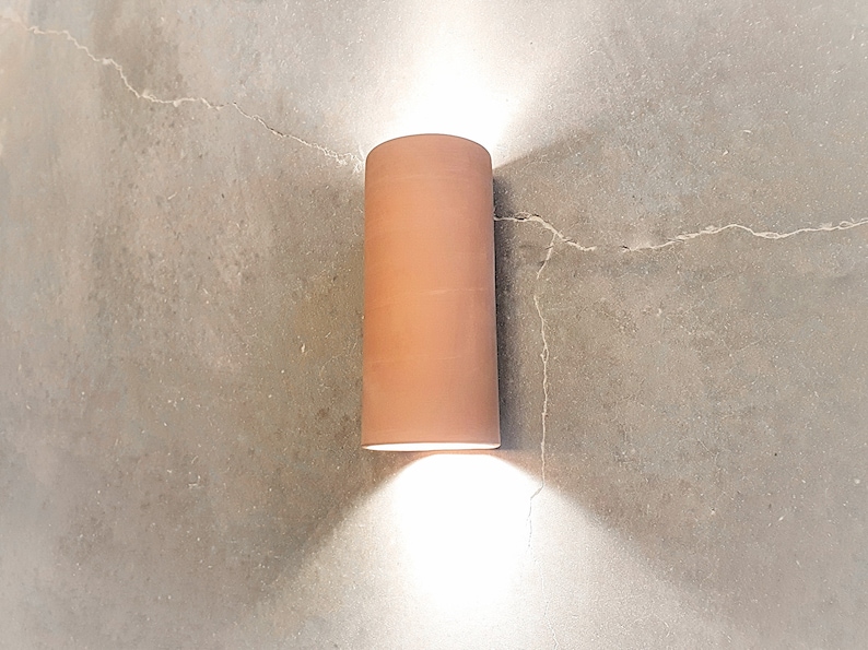 May include: A terracotta cylindrical wall sconce with a white light shining down on a concrete wall.