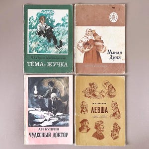 Puede incluir: Cuatro libros antiguos con cubiertas coloridas. Los libros presentan ilustraciones y texto en ruso. Los títulos incluyen "Tema y Zhuchka", "Smart Dunya", "Wonderful Doctor" y "Levsha". Los libros están dispuestos sobre una superficie neutra.