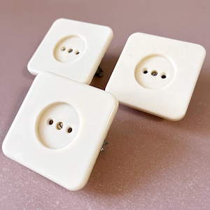 Puede incluir: Tres cubiertas de enchufes eléctricos vintage de color blanquecino. Cada cubierta cuadrada tiene un área circular empotrada con tres agujeros para las clavijas del enchufe. Las cubiertas son de un material brillante y liso y están montadas con clavijas metálicas.