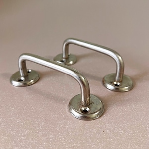 Könnte beinhalten: Zwei Vintage-Schrankgriffe aus silberfarbenem Metall. Jeder Griff hat einen gebogenen Griff, der an einer kreisförmigen Basis mit Schraubenlöchern befestigt ist. Die Griffe sind vor einem neutralen Hintergrund platziert.