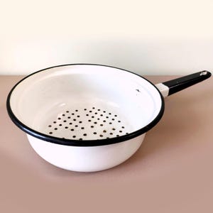 Puede incluir: Colador de esmalte blanco con borde y asa negros. El colador tiene pequeños agujeros en la base para drenar. El asa es negra y blanca. Una herramienta de cocina clásica para lavar y colar.