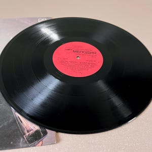 Puede incluir: Un disco de vinilo negro con una etiqueta roja con texto cir&iacute;lico. El disco est&aacute; colocado encima de una portada de &aacute;lbum parcialmente visible. La etiqueta dice "Melodiya" en escritura cir&iacute;lica. El disco es un art&iacute;culo de m&uacute;sica vintage.