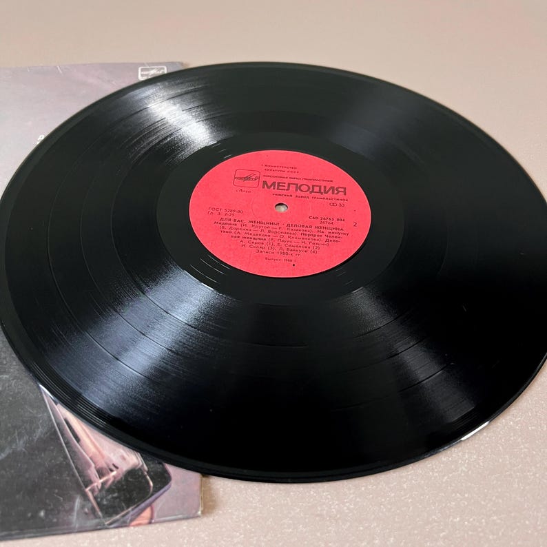 Puede incluir: Un disco de vinilo negro con una etiqueta roja con texto cir&iacute;lico. El disco est&aacute; colocado encima de una portada de &aacute;lbum parcialmente visible. La etiqueta dice "Melodiya" en escritura cir&iacute;lica. El disco es un art&iacute;culo de m&uacute;sica vintage.