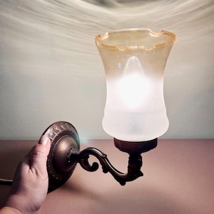 Puede incluir: Una lámpara de pared de estilo vintage con una pantalla de vidrio esmerilado y una base decorativa de metal color bronce. La pantalla tiene un borde con volantes y un cálido tono ámbar. La lámpara está diseñada para montarse en la pared y proporciona una iluminación ambiental suave.