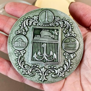 以下が含まれることがあります： 丸い緑と銀色の金属メダルで、装飾的なディテールが施されています。中央には、錨、船、星の盾があります。メダルには、建物と灯台のデザインが施された3つの円形エンブレムがあります。