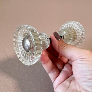 Può includere: Una coppia di pomelli per porte in vetro trasparente con un design sfaccettato. I pomelli sono collegati da un'asta di metallo.