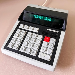 Peut inclure: Une calculatrice vintage avec un corps blanc et noir, affichant les chiffres 1234567890 sur un écran numérique vert. La calculatrice comporte de nombreux boutons, dont un grand bouton rouge "C". Le nom du modèle "ЭЛЕКТРОНИНА МН-44" est visible.