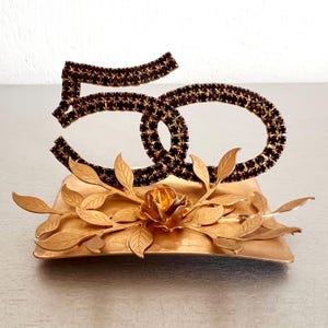 Peut inclure: Centre de table en métal doré avec le chiffre "50" incrusté de strass foncés. Le chiffre est posé sur une base carrée dorée ornée d'une rose et de feuilles. Un objet décoratif pour une célébration importante.