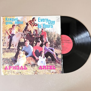 Peut inclure: Un disque vinyle vintage avec un label noir et une pochette colorée représentant un groupe appelé "Ariel Group" avec le texte "Every Day Is Yours" et "Каждый день твой" en russe. La pochette montre les membres du groupe avec des chevaux et un chien dans un champ.