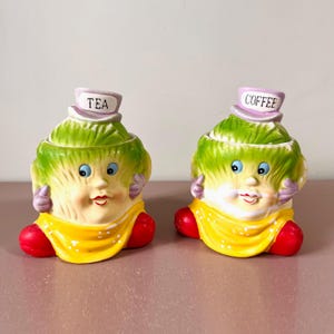 Puede incluir: Dos botes de cerámica caprichosos, uno con la etiqueta "TEA" y el otro "COFFEE". Cada bote tiene forma de cara sonriente con una cabeza verde parecida a una lechuga, un babero amarillo y pies rojos. Diseño vintage.