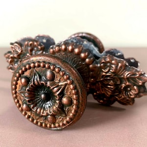 Peut inclure: Un ensemble de trois boutons de porte ornés de couleur cuivre avec des motifs floraux complexes. Les boutons ont un bord surélevé perlé et un motif floral central.