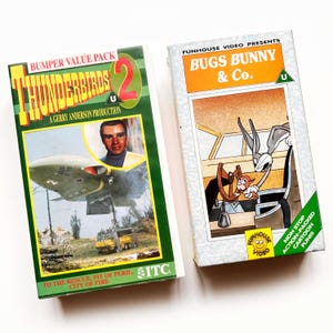 Può includere: Due videocassette VHS vintage. Una presenta "Thunderbirds 2" con una copertina verde e gialla, e l'altra è "Bugs Bunny & Co." con Bugs Bunny e un personaggio dei cartoni animati. Entrambe le cassette sono nella loro confezione originale.
