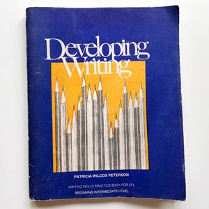 Può includere: Un libro vintage intitolato "Developing Writing" con una copertina blu e un rettangolo arancione al centro. Il titolo è in bianco, con una grafica di matite. Il libro è un quaderno di esercizi di scrittura per EFL, livello intermedio principiante.