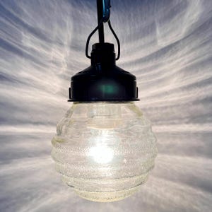 Op de afbeelding: Een heldere glazen hanglamp met een getextureerd, geribbeld ontwerp. De lamp heeft een zwarte dop en een metalen haak om op te hangen. De gloeilamp binnenin is verlicht en geeft een warme gloed.