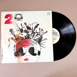 May include: A vintage vinyl record album with a white cover featuring a stylized illustration of a face wearing sunglasses and a headband with Russian text. Musical instruments are depicted above the face. A black vinyl record is next to the album.