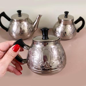 May include: Three silver-colored teapots with black handles and lids. The teapots have an ornate, embossed floral design. One teapot has a spout, while the other two have rounded bodies. The handle of one teapot is being held by a person.
