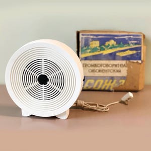 May include: A white, circular vintage speaker with a concentric grill design. The speaker has a black center and is connected to a power cord. A cardboard box with faded blue and yellow graphics is in the background.